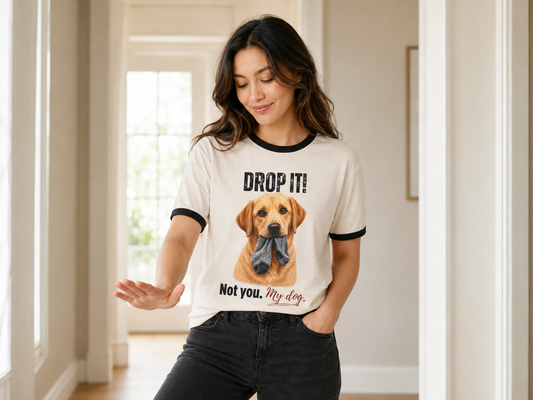 Woman wearing a cream ringer tee with black trim, printed with "Drop It! Not you. My dog." and a hand-illustrated yellow Labrador holding a grey sock, standing in a sunlit hallway with one hand raised