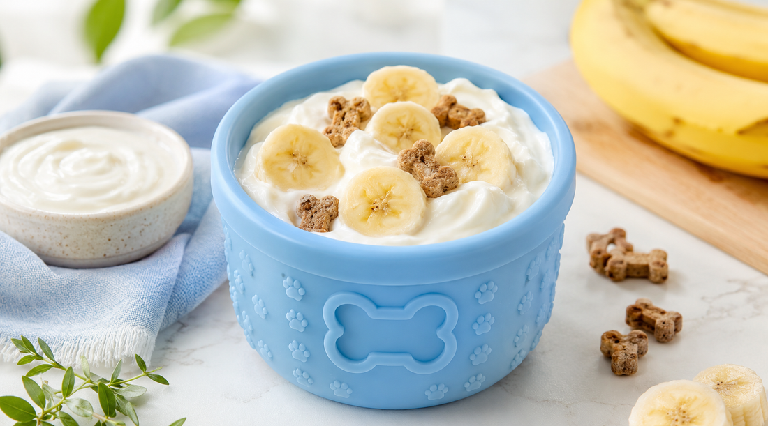 Blue silicone dog lick cup with a paw-print and bone design, filled with whipped yogurt, banana slices, and dog treat pieces, styled next to a bowl of yogurt, a blue cloth, greenery, and fresh bananas