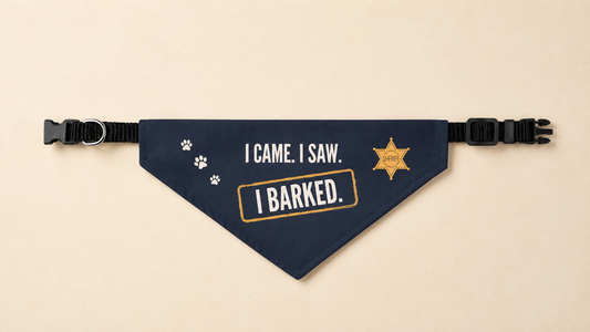 Navy blue dog bandana with white paw prints and a mustard-gold sheriff badge reading "SHERIFF," printed with "I CAME. I SAW." and a stamped "I BARKED." box, clipped onto a black adjustable collar with a silver D-ring, laid flat on a cream background