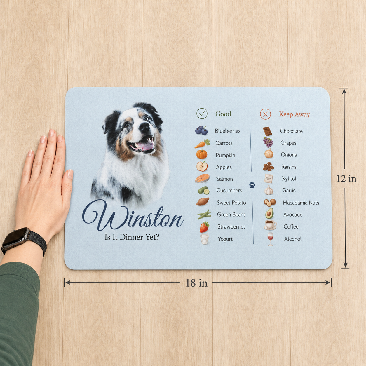 Hand beside the 12 x 18 inch personalized dog food mat, showing its size against a wood floor.