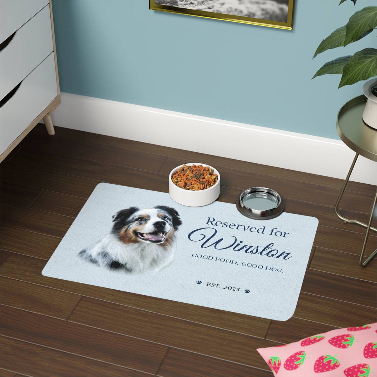 Blue personalized dog placemat in a feeding corner on dark wood, with a kibble bowl and a water bowl.