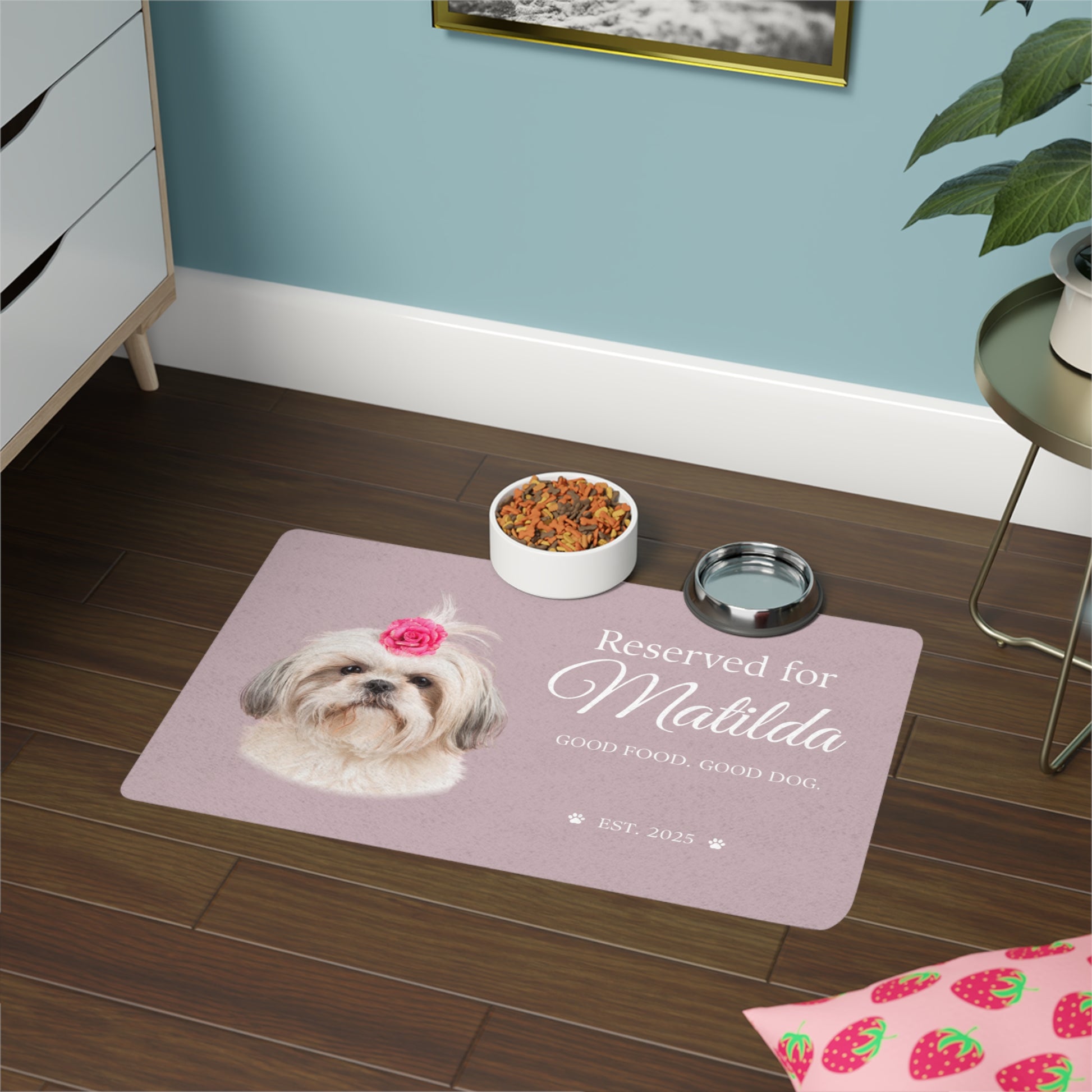 Rose dog placemat lettered Reserved for Matilda on a dark wood floor beside a dresser and two bowls.