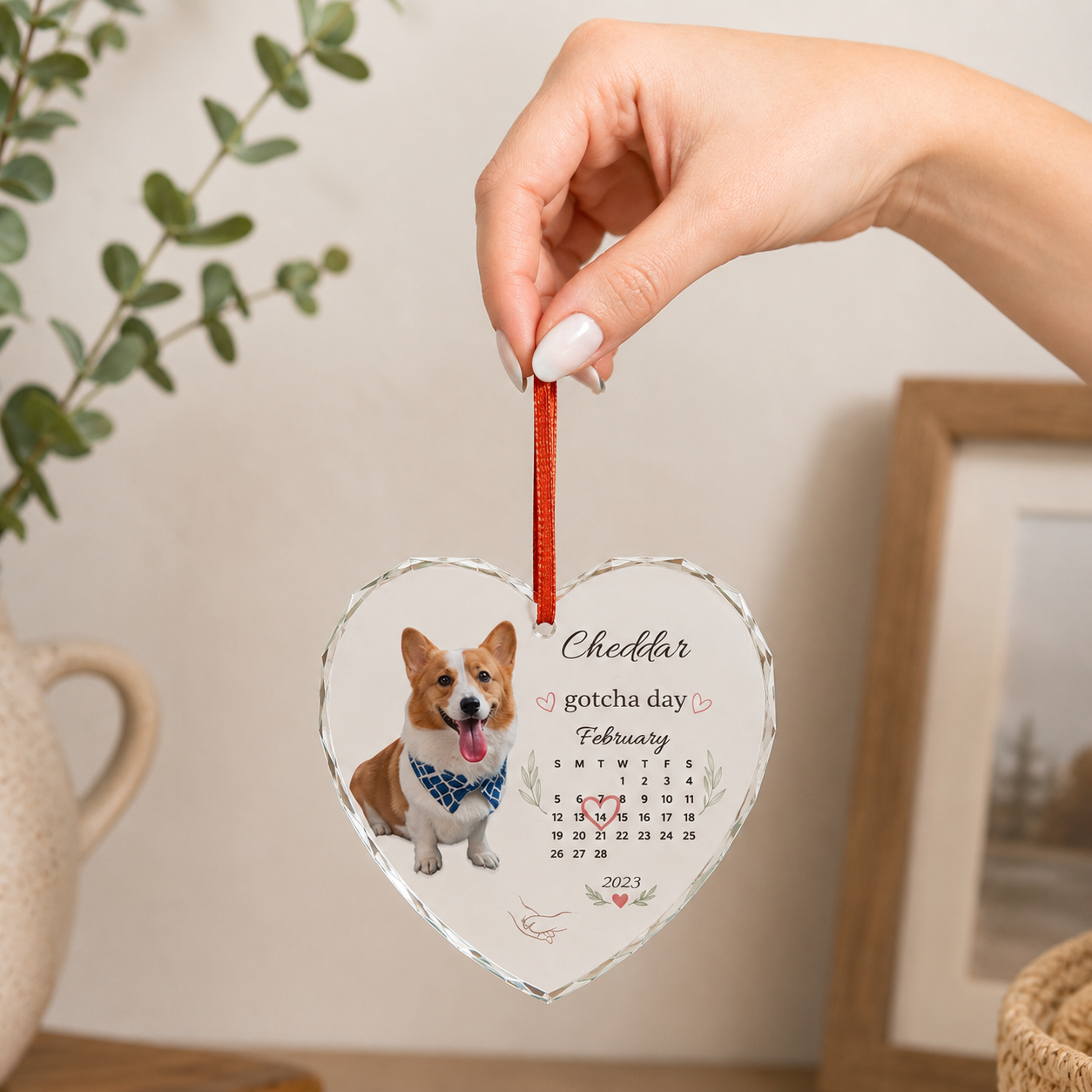 Hand holding a personalized gotcha day dog ornament by its red ribbon near a potted plant.