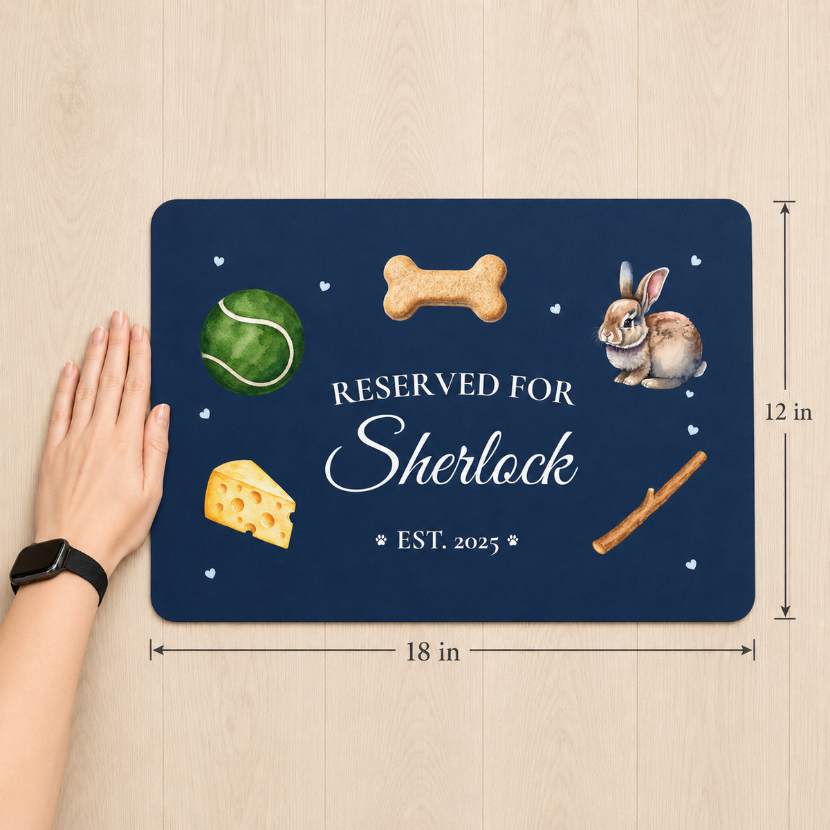 Navy blue personalized dog placemat that reads 'Reserved for Sherlock, Est. 2025' with dog toy illustrations — tennis ball, bone, cheese wedge, rabbit toy, and stick — shown at 12 x 18 inches next to a hand for size reference.