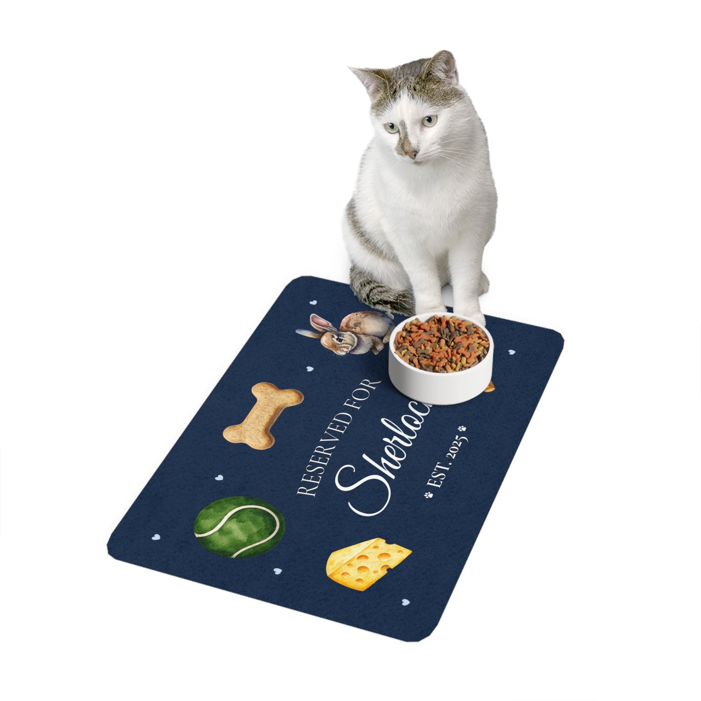 White and grey cat sitting on a navy personalized pet feeding mat next to a bowl of food.